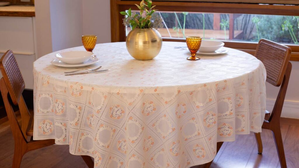 Imagem que mostra uma mesa redonda com uma toalha de mesa redonda acima dela. Sobre a mesa está um vaso de plantas dourado com plantas dentro e, dispostas em diferentes pontos da mesa estão pratos, talheres e dois copos