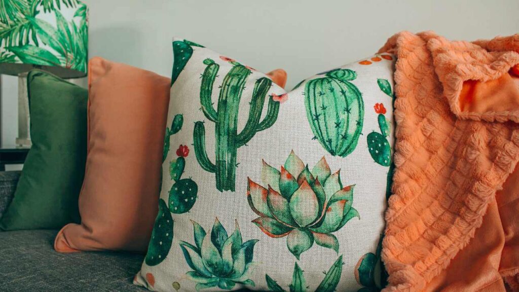 Um sofá cinza e, acima dele, estão três almofadas. A almofada no canto esquerdo é verde, a do centro é laranja e ao lado é branca com estampas de cactos. Ao lado das almofadas está um tecido laranja