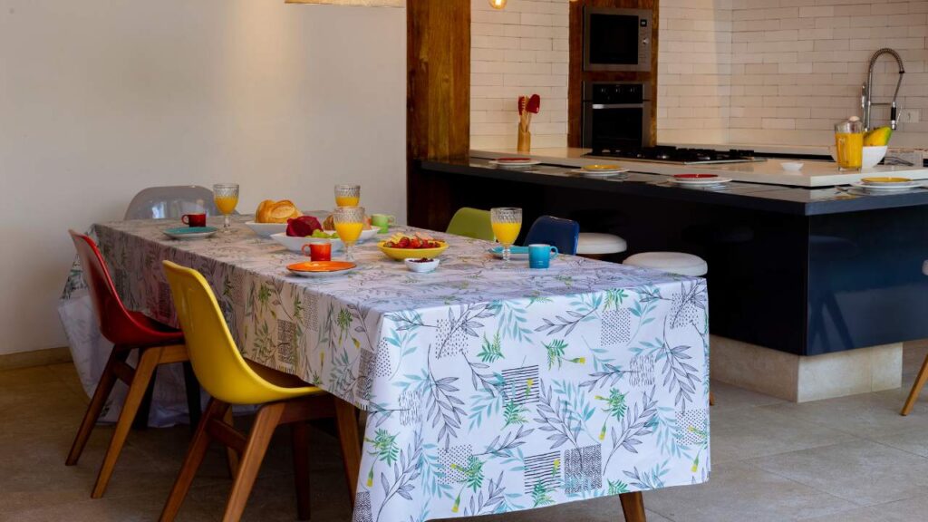 Uma mesa com uma toalha de mesa estampada. Acima, estão pratos, jarras de suco e xícaras
