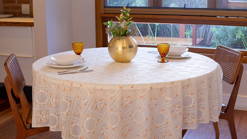 Mesa redonda de jantar com toalha branca bordada, adornada com um vaso dourado e dois copos de vidro âmbar.