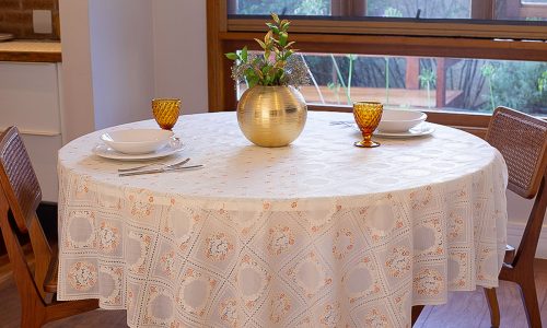 Mesa redonda de jantar com toalha branca bordada, adornada com um vaso dourado e dois copos de vidro âmbar.